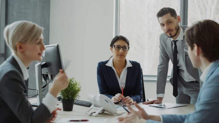 Business professionals collaborating around a table in a modern office, with a businesswoman in a navy blazer and glasses holding a pencil while speaking with male and female colleagues