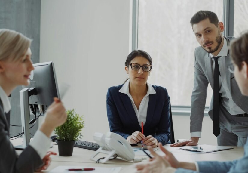 Business professionals collaborating around a table in a modern office, with a businesswoman in a navy blazer and glasses holding a pencil while speaking with male and female colleagues