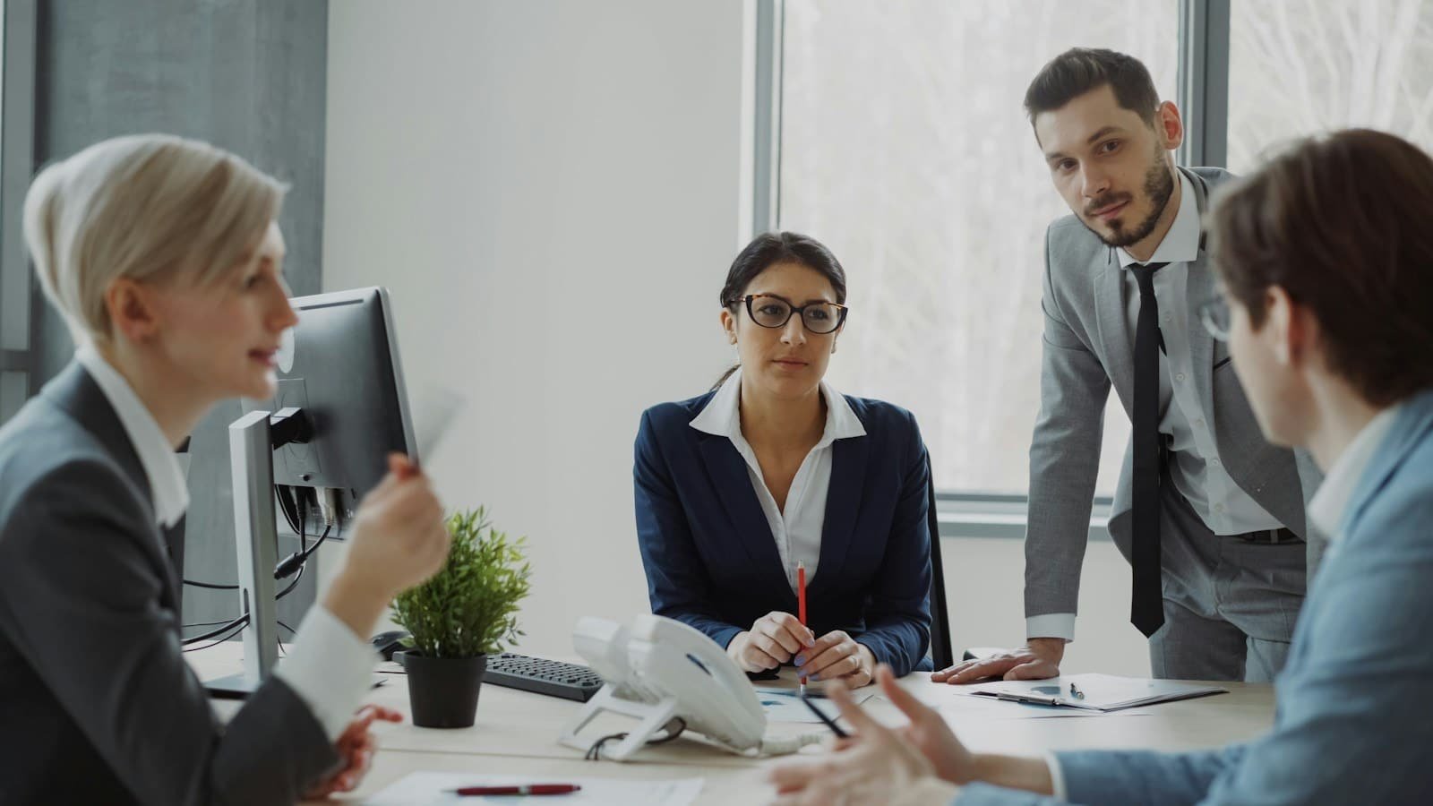 Business professionals collaborating around a table in a modern office, with a businesswoman in a navy blazer and glasses holding a pencil while speaking with male and female colleagues