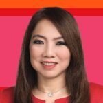 A woman with long brown hair wearing a red jacket and a silver flower necklace, smiling, photographed against a pink and orange graphic background