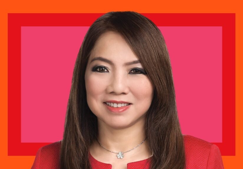 A woman with long brown hair wearing a red jacket and a silver flower necklace, smiling, photographed against a pink and orange graphic background