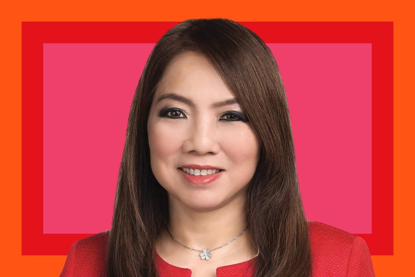 A woman with long brown hair wearing a red jacket and a silver flower necklace, smiling, photographed against a pink and orange graphic background